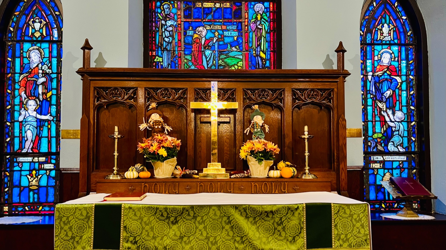 Church altar decorated with autumn flowers and a brass cross, framed by stained-glass windows depicting St. Mary on the left, St. Alban in the center, and St. Dorcas on the right.