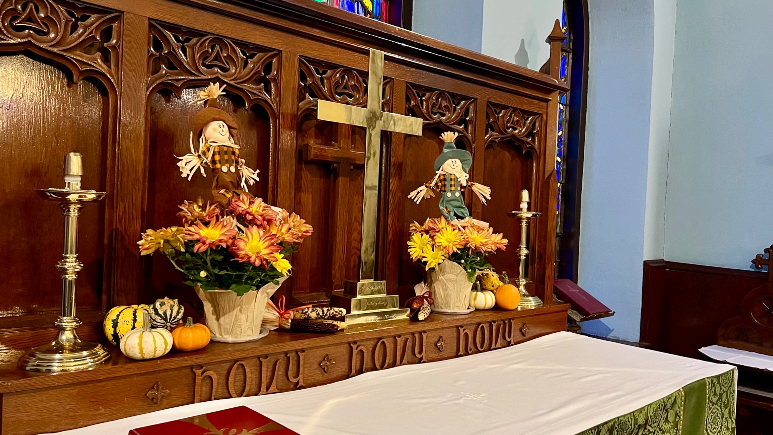 Autumn-decorated church altar with brass cross, candles, colorful chrysanthemums, small pumpkins, and cheerful scarecrow figures arranged before a carved wooden reredos.