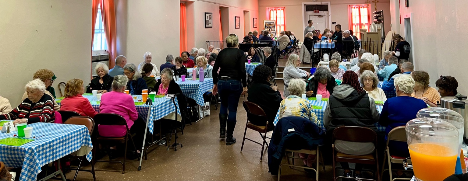 Large group gathered in a church hall for a community meal, seated at long tables with blue checkered tablecloths, sharing food and conversation in a warm, lively atmosphere.